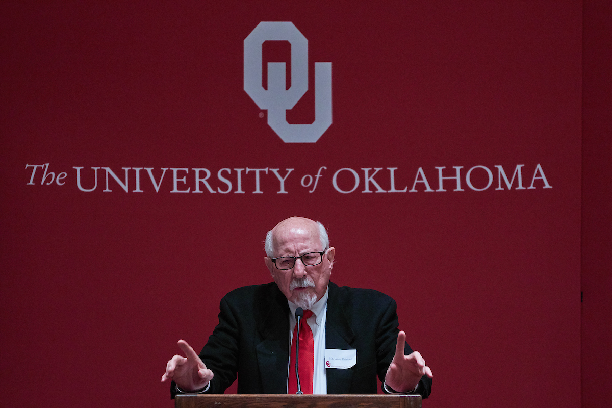 Gene Rainbolt speaking at a podium in front of a red background that reads "OU. The University of Oklahoma."