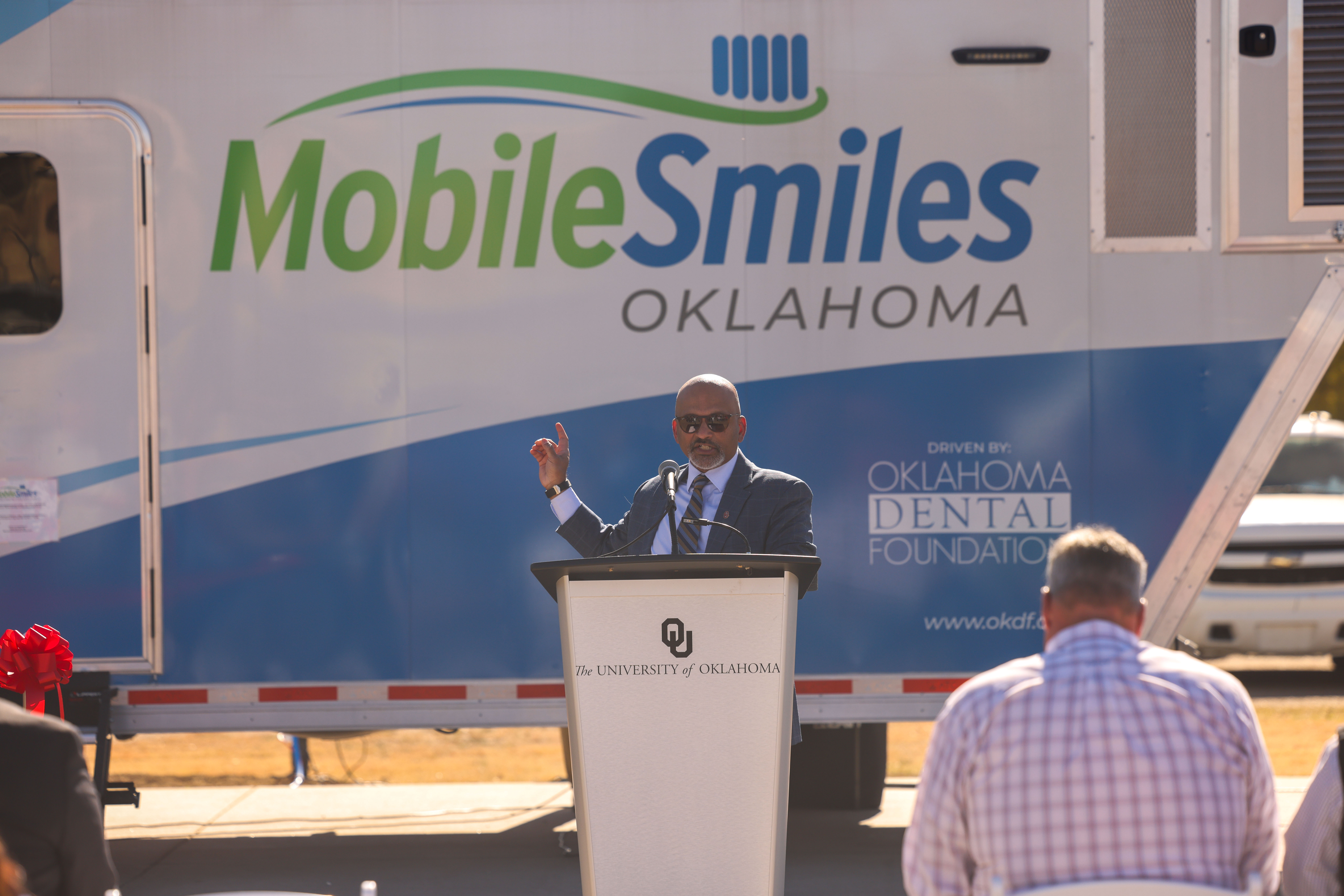 A man stands at a podium in front of the Mobile Smiles Oklahoma clinic, pointing at it. 