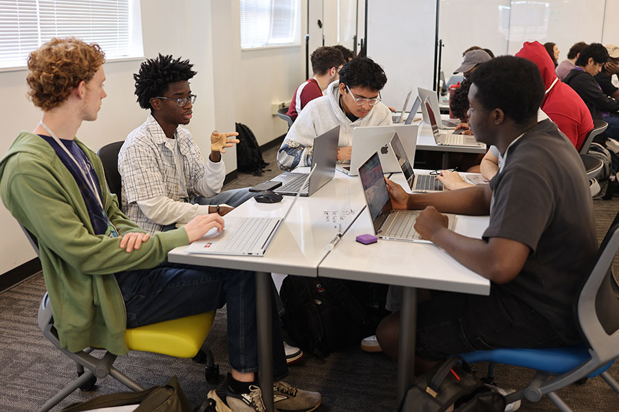 A group of students sit together at a table, working on laptops and talking.