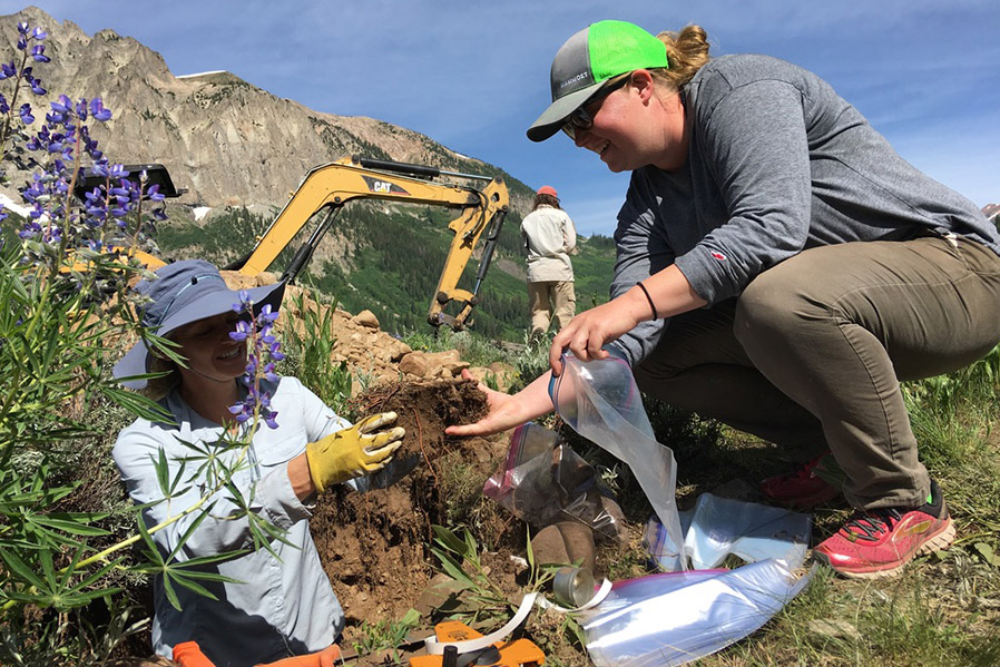 Two people harvest soil from a lab site. One stands in a hole in the ground, handing a clump of dirt to a person standing at the surface.