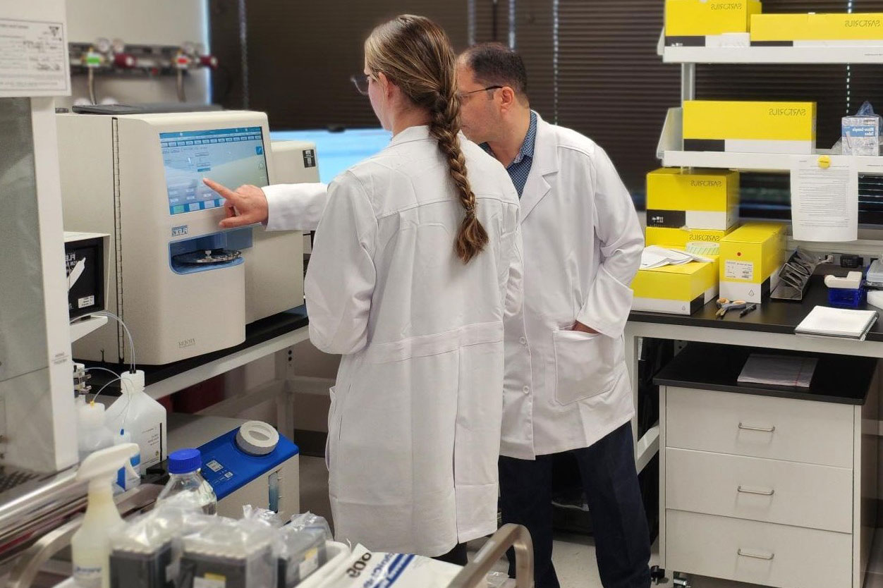 Two researchers in white coats stand in a laboratory setting, looking at a computer screen.