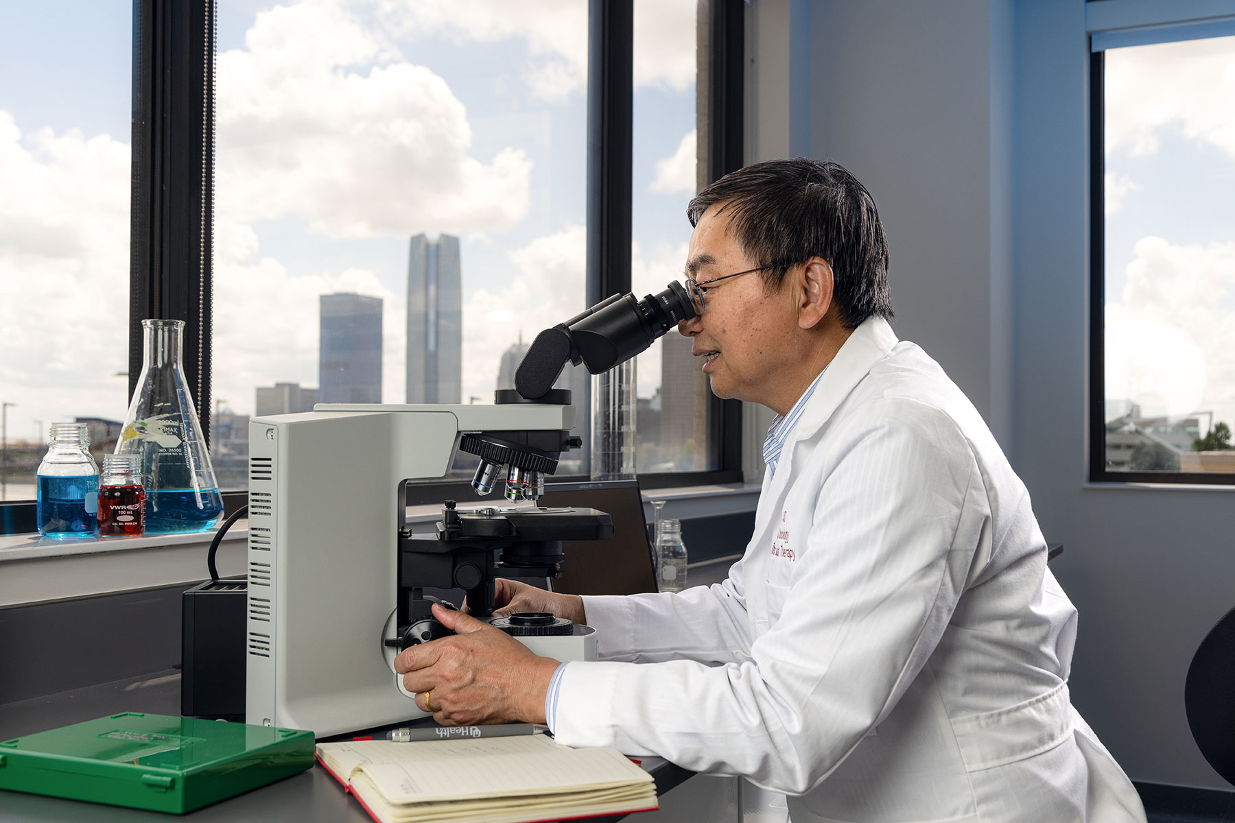 Yubin Kang looks into a microscope at a desk seated in front of a window. Outside the window is the Oklahoma City skyline.