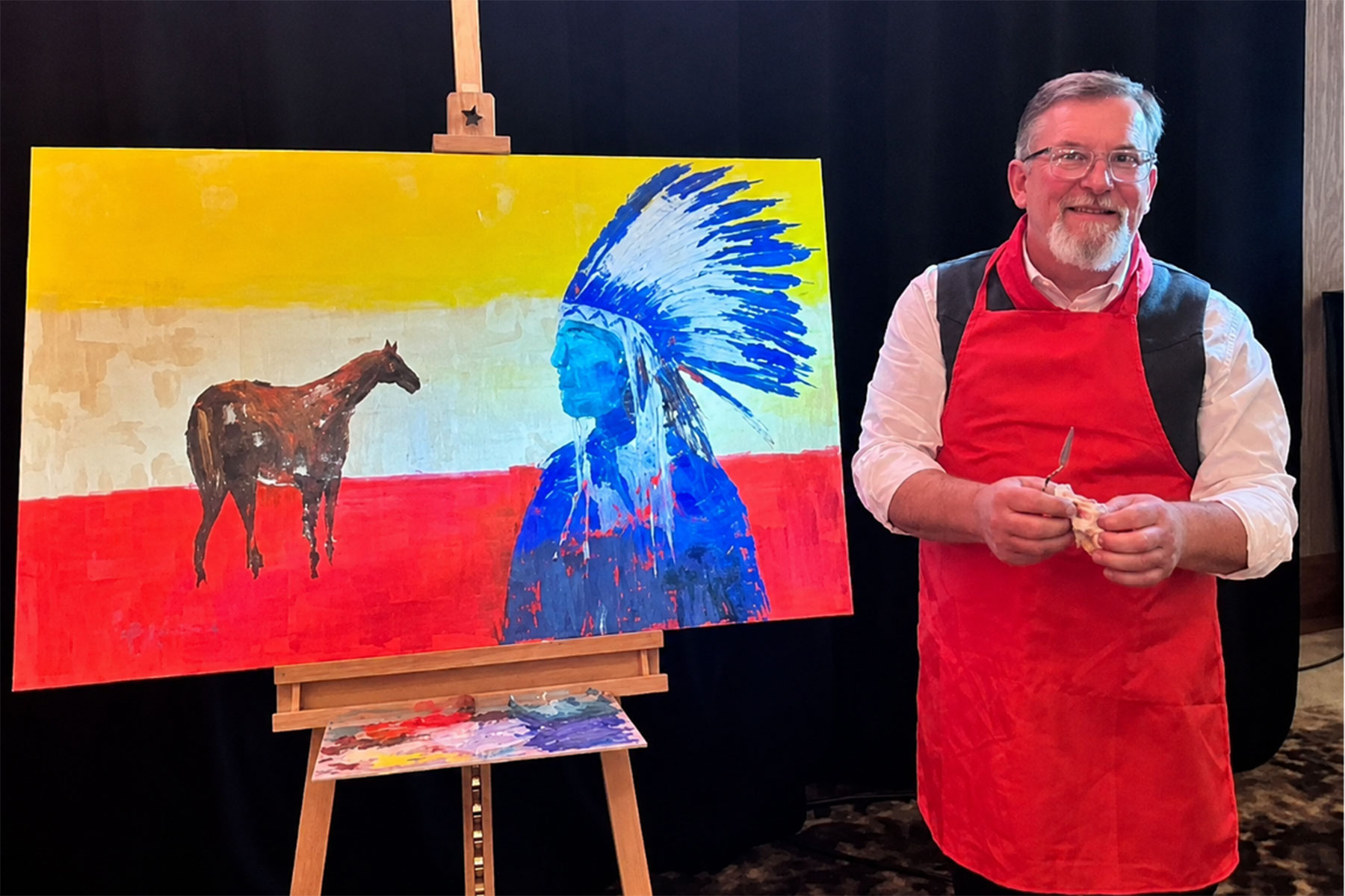 A man stands next to a painting of a horse and an indigenous person in traditional headdress.