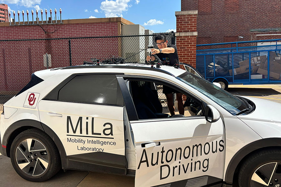 A white sedan with the Words "MiLa Mobility Intelligence Laboratory" on the back door and "Autonomous Driving" on the front door. A person stands beside the vehicle, fitting equipment to the roof.