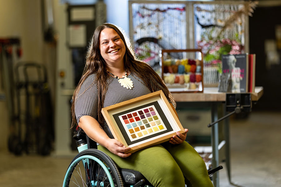 A woman seated in a wheel chair holds up a piece of art in a frame.