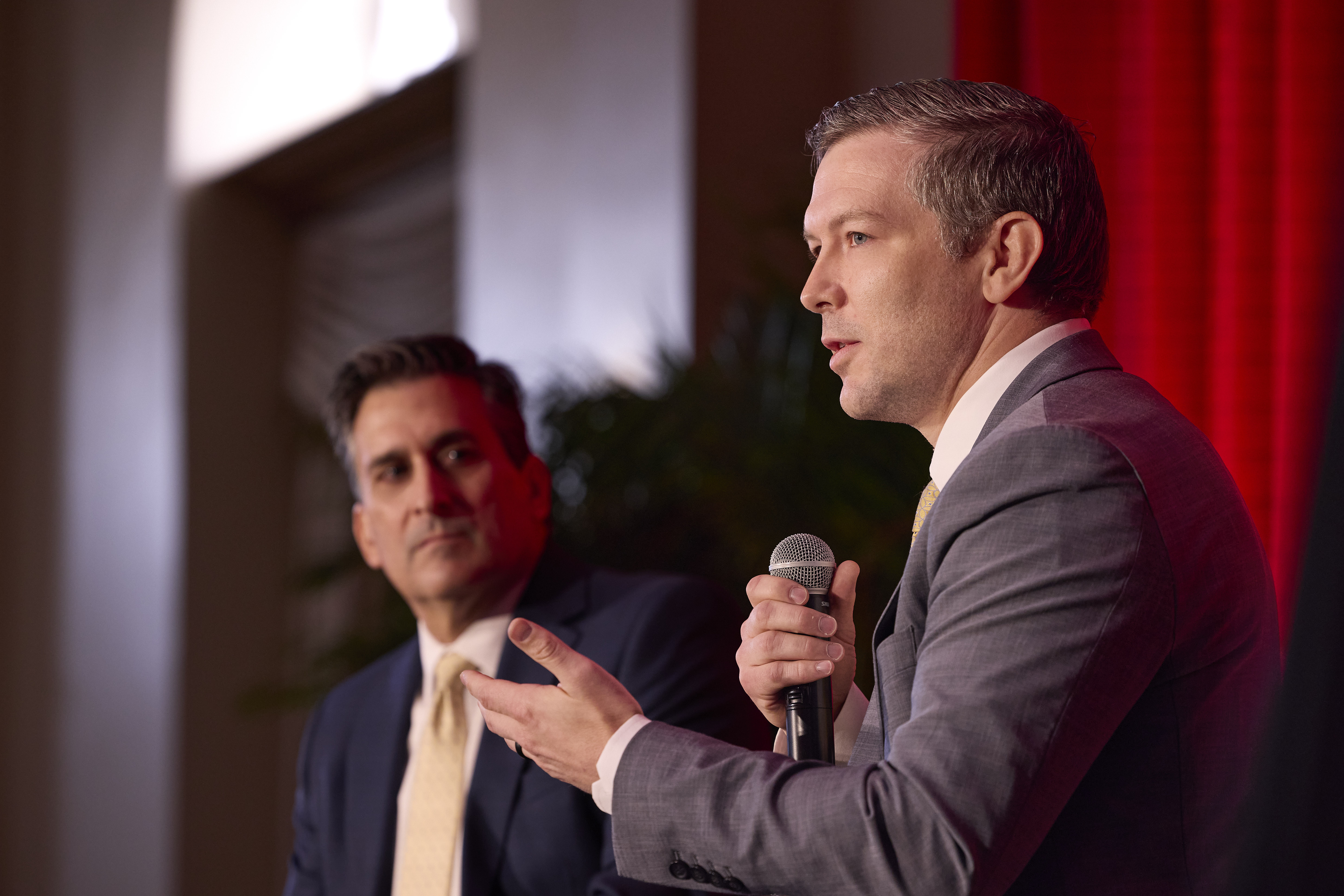 Two men sitting on a stage. The man in the foreground is holding a microphone and speaking.