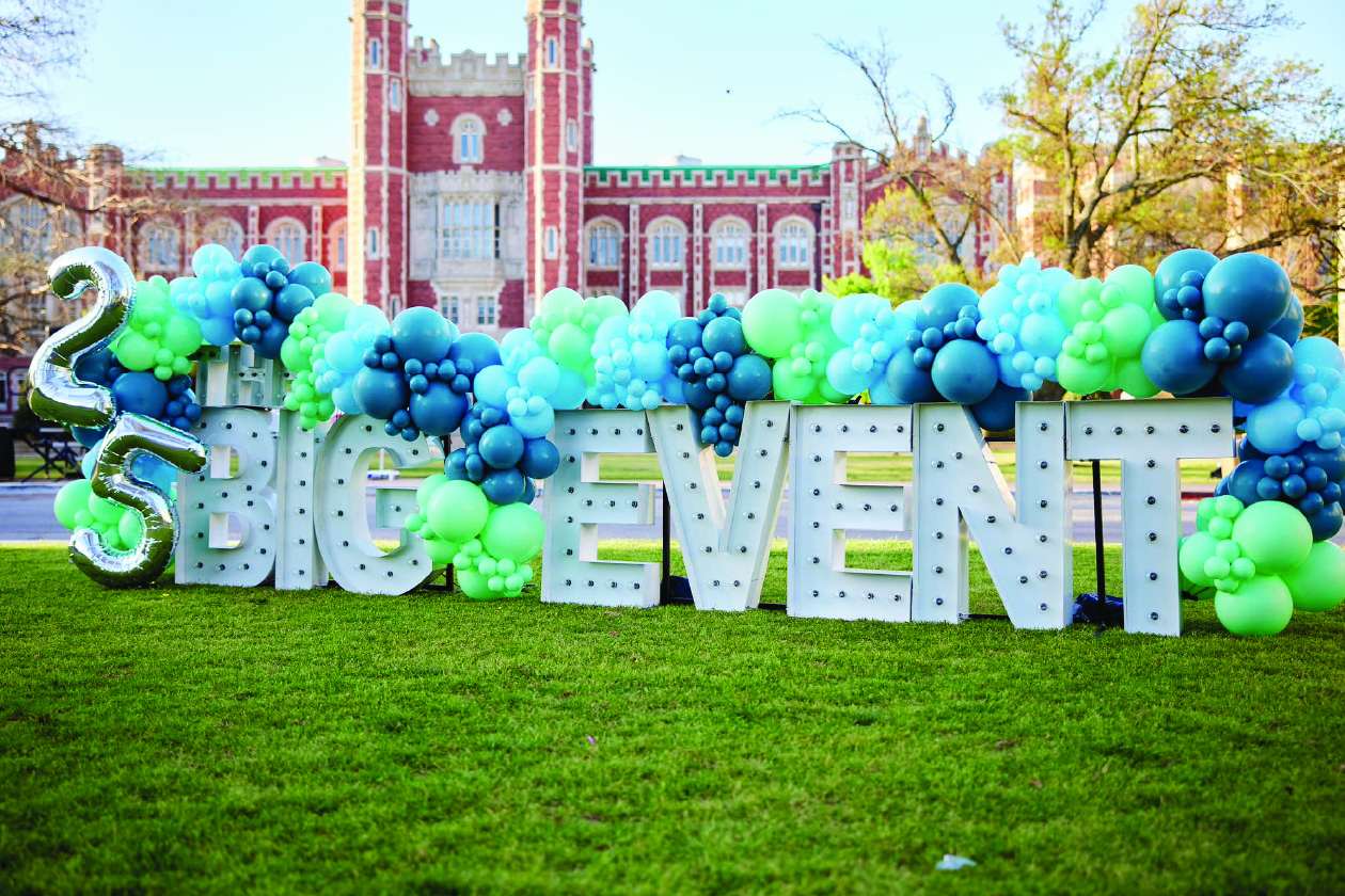 25 in silver balloons, followed by Big Event in giant letters, surrounded by additional balloons, with buildings and trees in the background.
