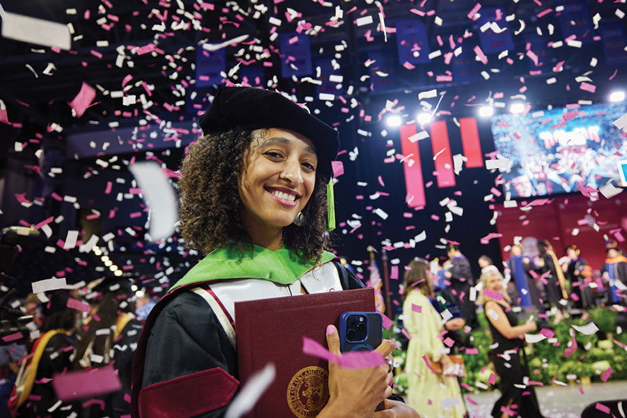 A student in graduation regalia, with confetti and lights in the background.