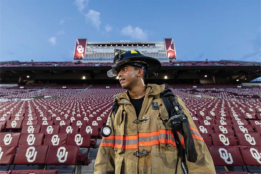 A cadet in full firefighter gear standing in front of the seats of Gaylord Family Memorial Stadium.