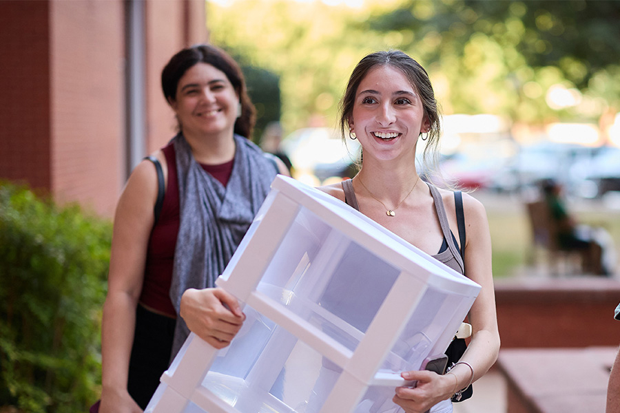 A new student bringing in a white storage container with another new student in the background smiling.