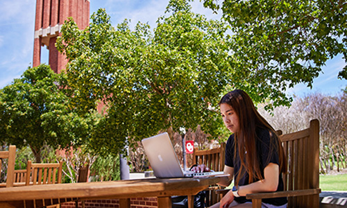 Female OU student sitting at an outside table under blue sky while working on her computer