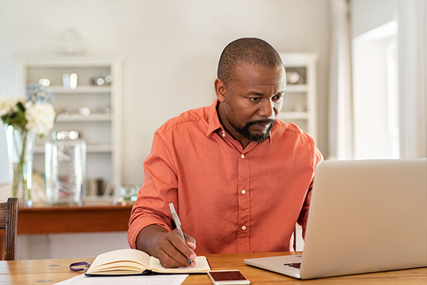 Man taking notes while looking at a laptop.