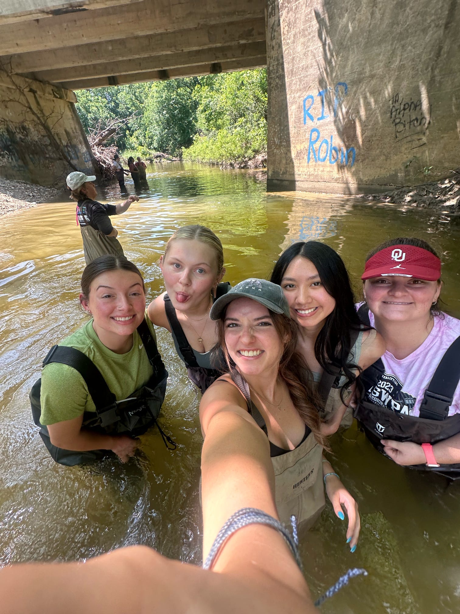 Students wearing waders stand together in shallow creek water during a field sampling activity as part of a summer biological station experience.