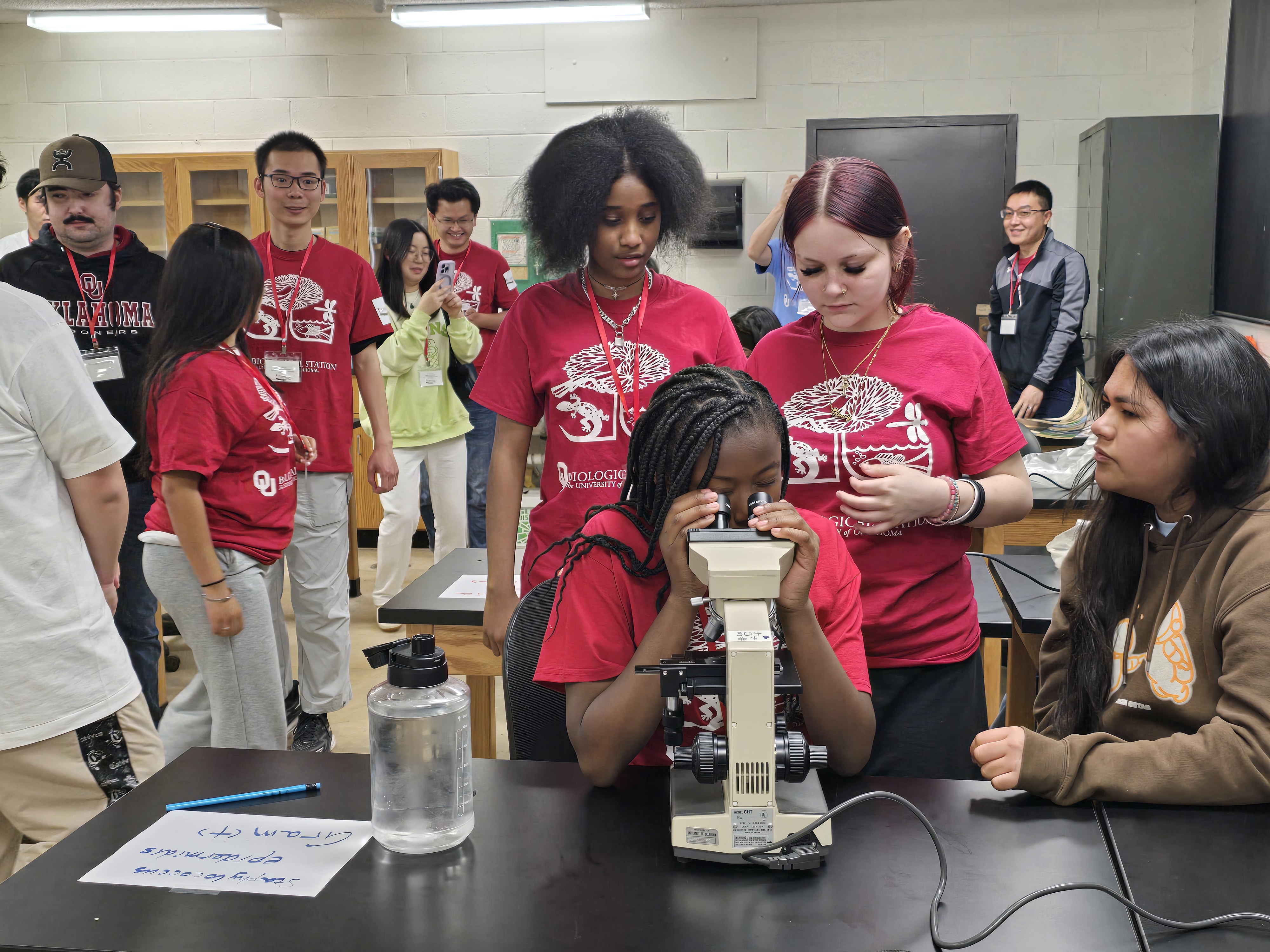 Students gathered around a microscope in a biology classroom as one student observes a sample while others watch and discuss during a hands-on laboratory activity.