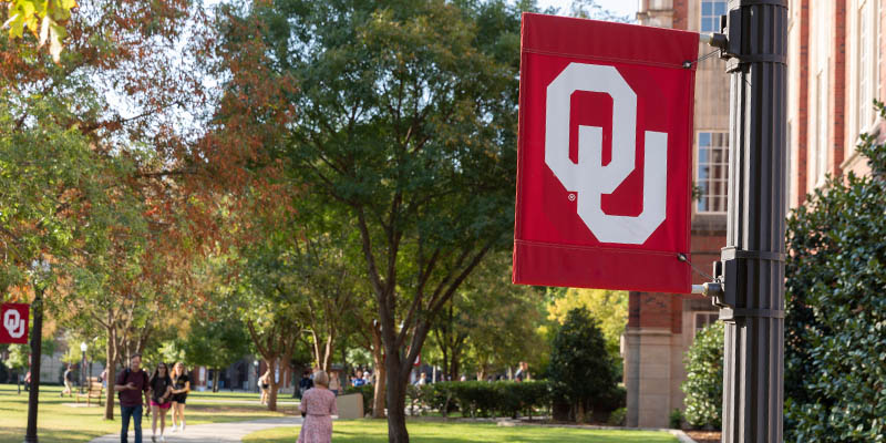 close-up of a crimson OU flag mounted on a lampost and students on campus walking to class 