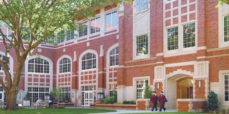 exterior of Price Hall with two students dressed in cap and gown walking across the courtyard