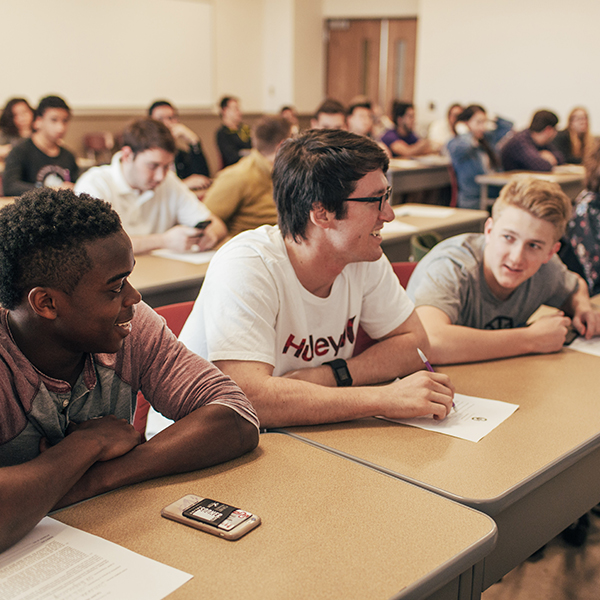 A classroom with students seated at small individual desks, each with either papers or phones on their desks. In the first row, three students are smiling and talking to each other. The student in the middle is writing something on a piece of paper