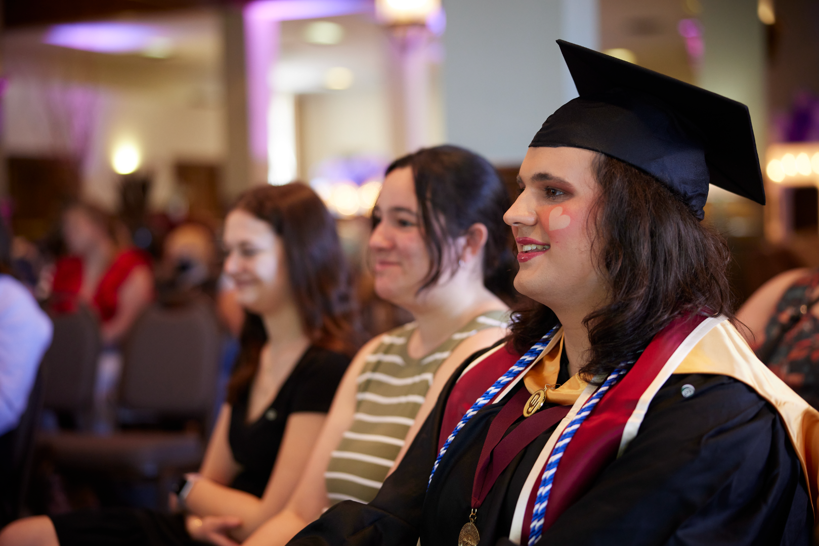 Students in graduation regalia, with rainbow cords and a heart tattoo on their face, room is lit up in Lavender lights