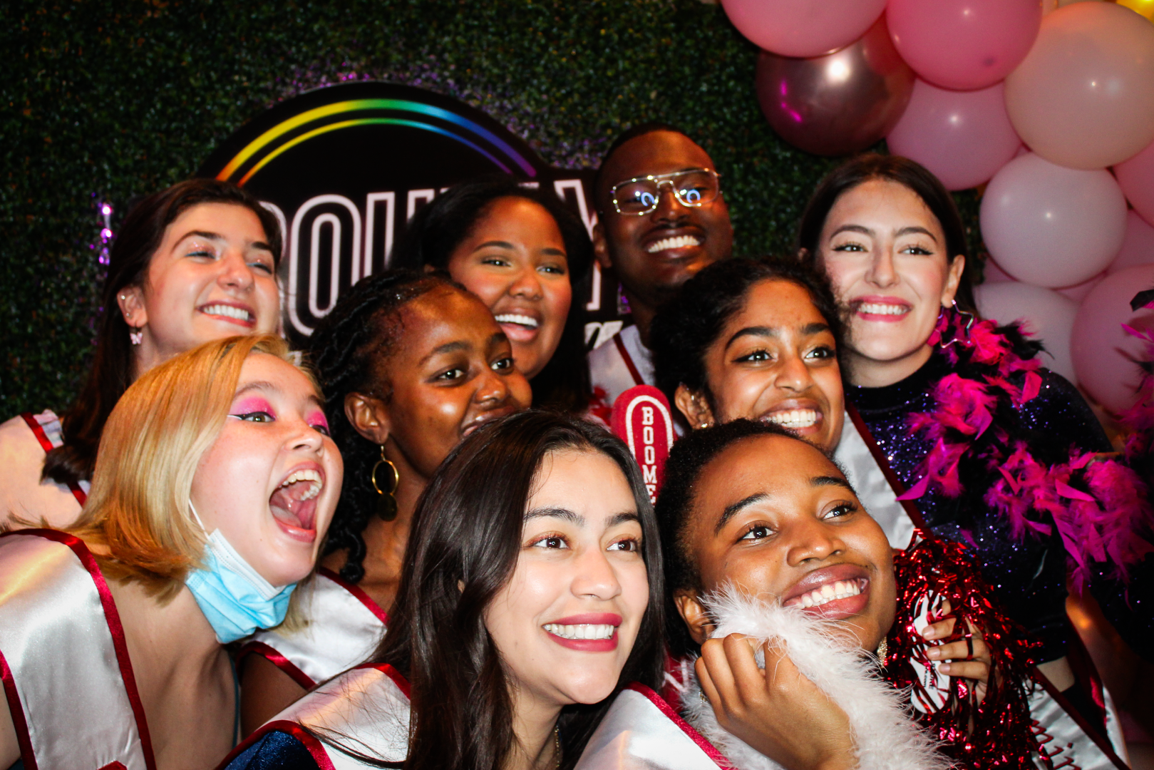 A group of students smiling and taking a group photo in front of a grass wall with pink balloons