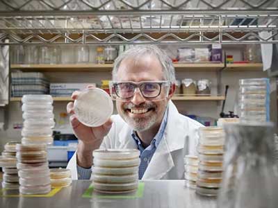 A man in a scientific labcoat smiles holding a petree dish.