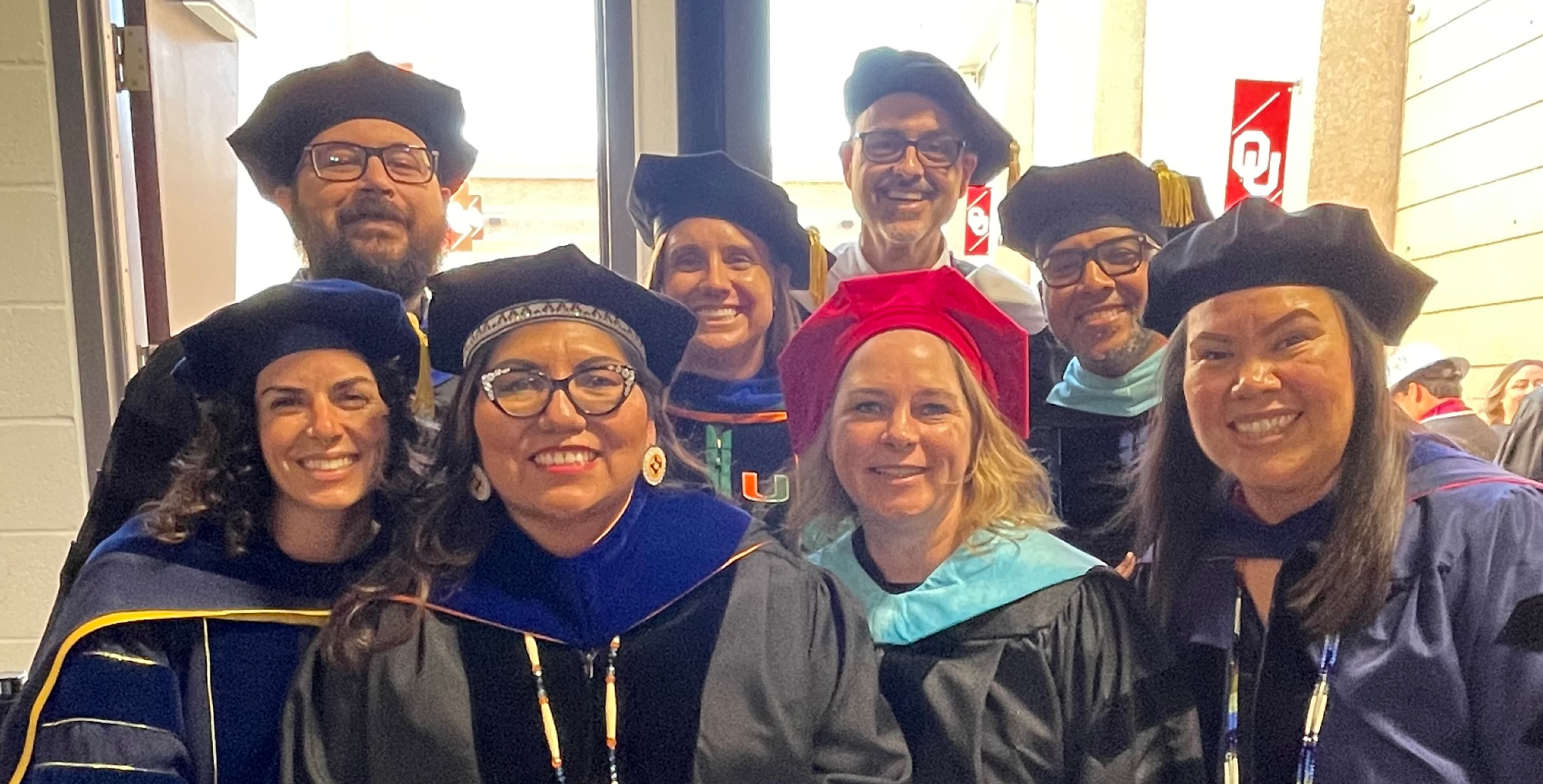 Students and faculty in graduation regalia standing in front of a Jeannine Rainbolt College of Education banner.