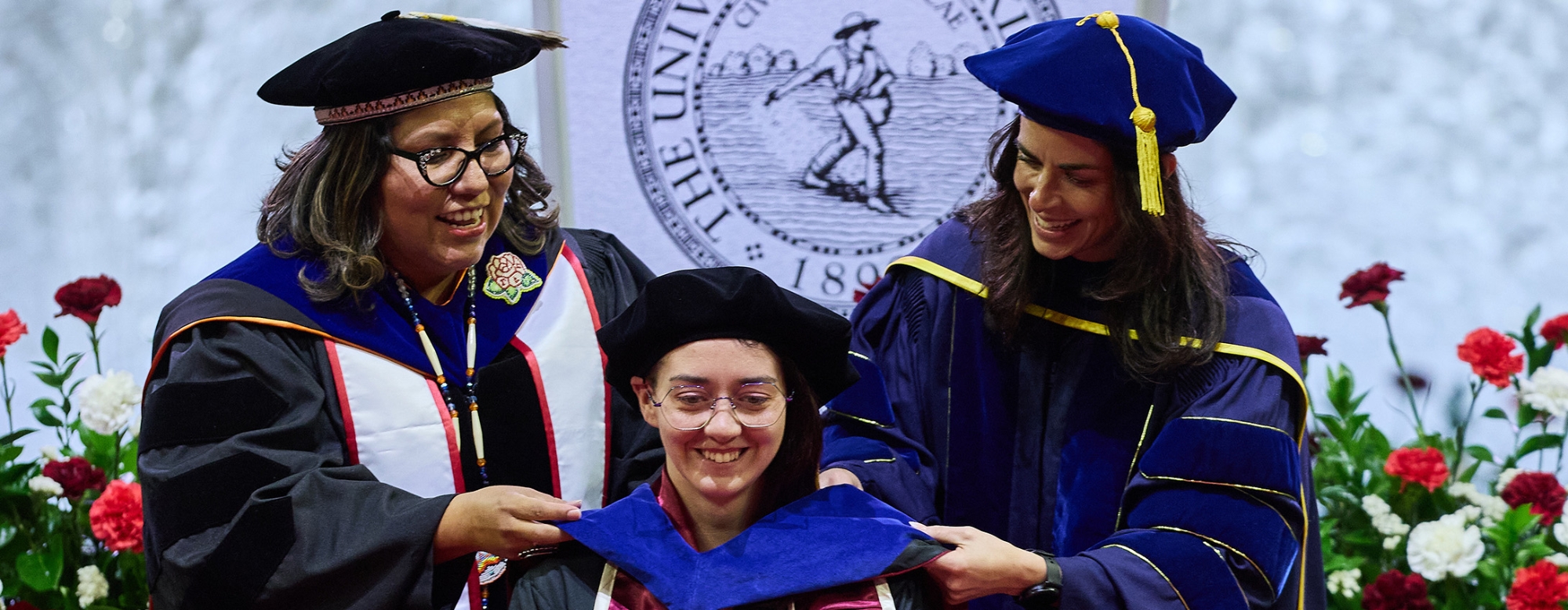 Students and faculty in graduation regalia standing in front of a Jeannine Rainbolt College of Education banner.