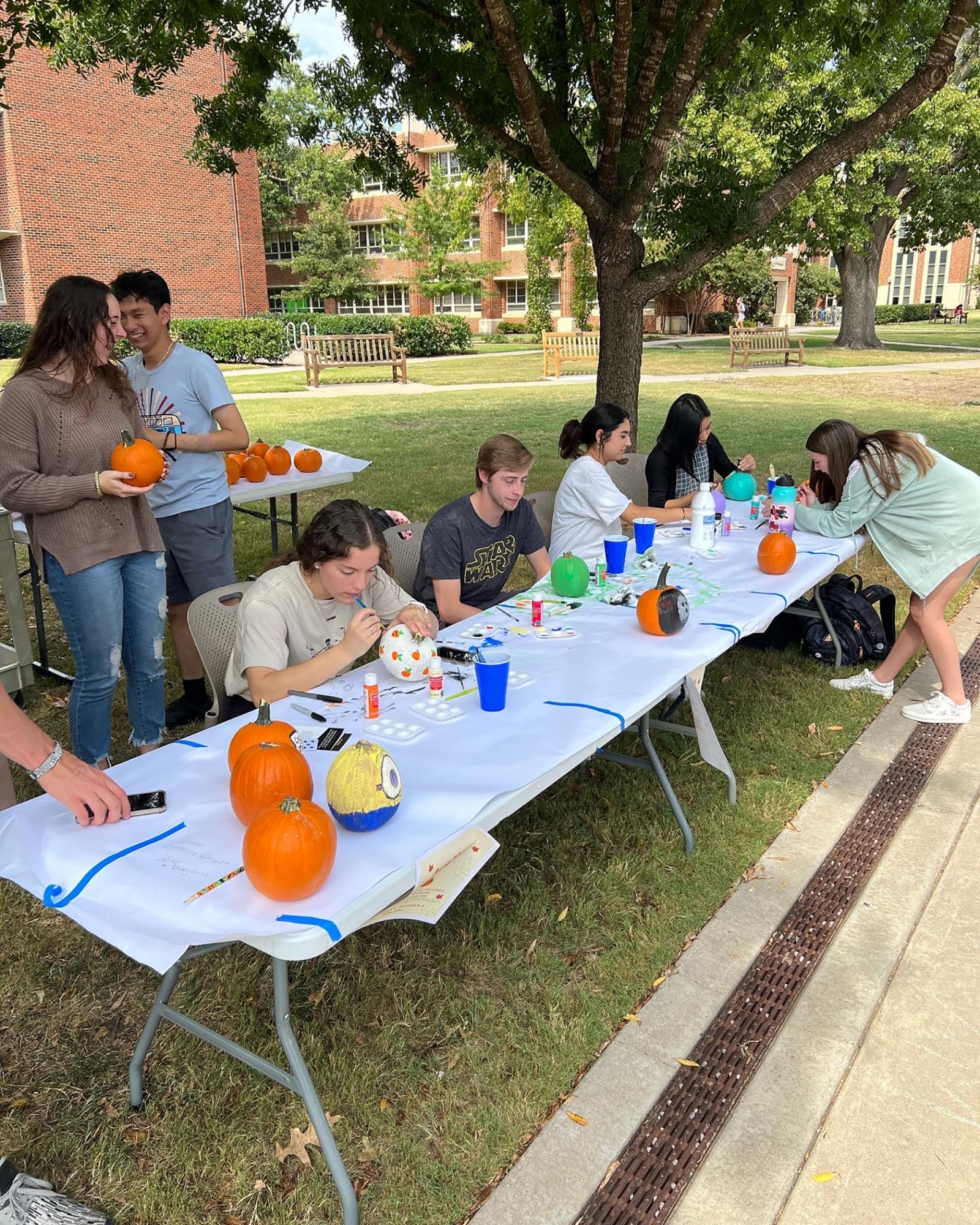 students sitting at a table under a tree painting pumpkins