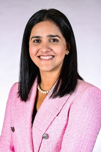 woman with dark hair wearing a pink blazer and pearls smiling for a photo