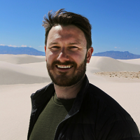 a man in a dark jacket posing for a picture outside in front of sand dunes