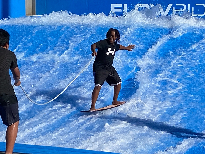 Student wearing black shorts and a black shirt on a surfboard holding a rope