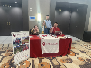 Four students posing behind a table with OU swag in the lobby of a conference center