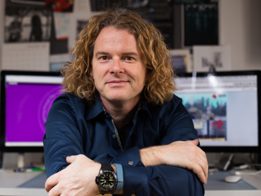 Man in blue shirt with wavy hair sitting in front of two computer screens posing for a photo