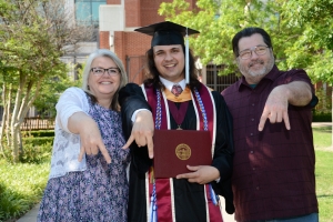 student with two family members holding diploma cover and giving a horns down