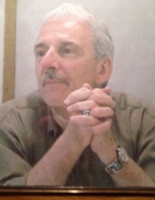 Man in brown shirt sitting with his hands clasped by his chin