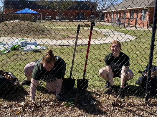 Two students sitting behind a chainlink fence digging in the dirt