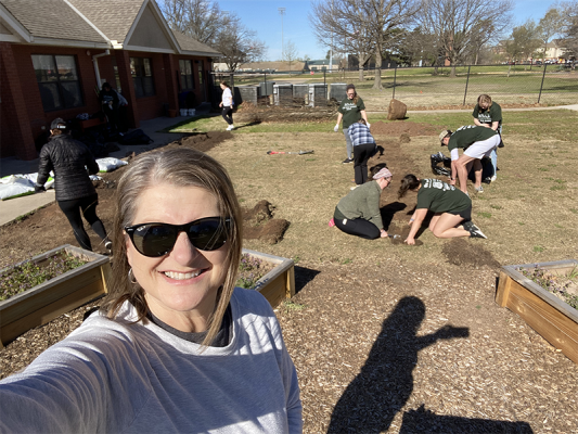 A woman in a white shirt outside taking a selfie with students planting in the background