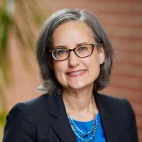 Woman with grayish hair and glasses wearing a navy blazer and blue blouse