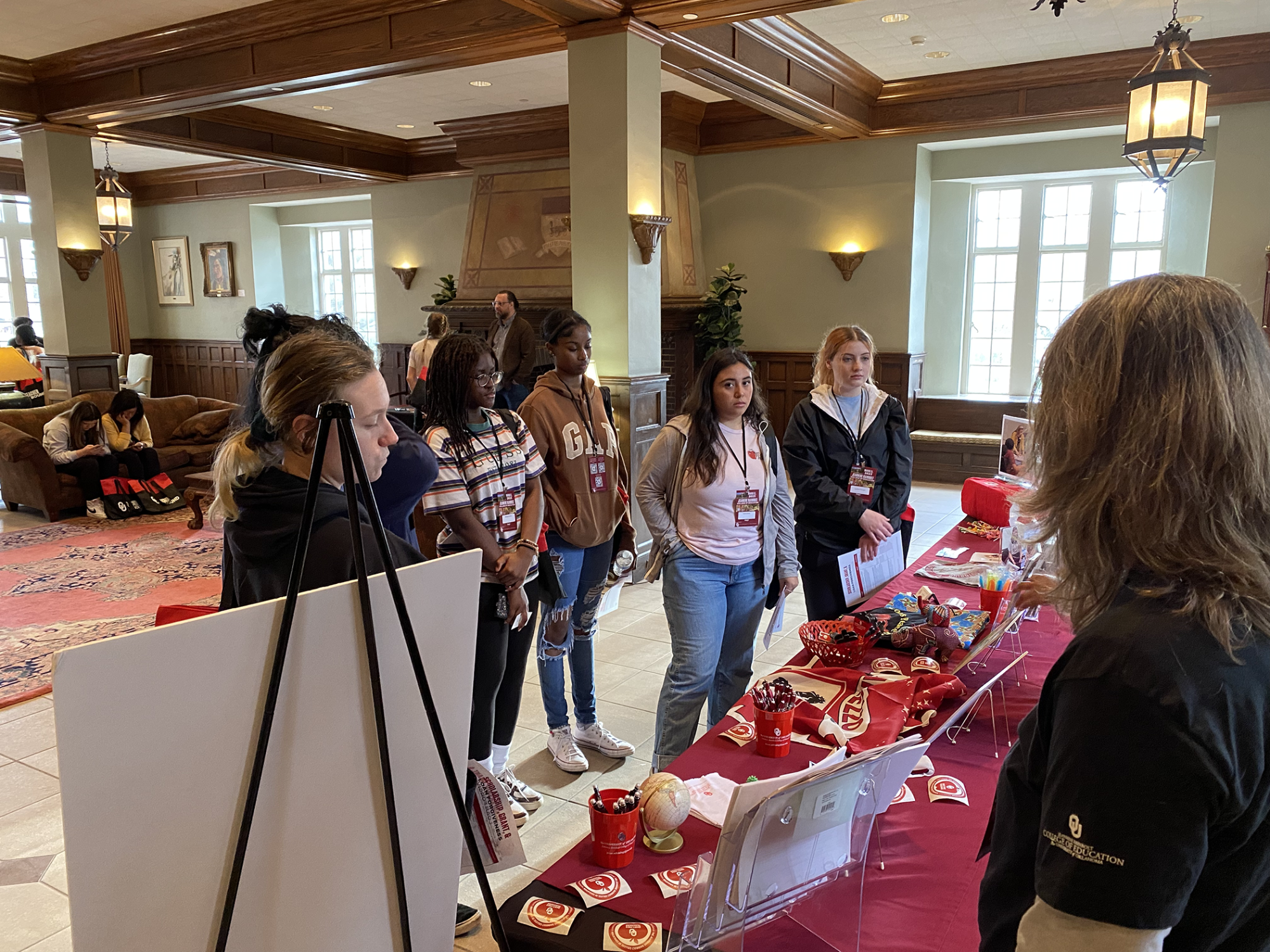 Students in an open room standing in front of a table with a crimson table cloth while a woman speaks to them