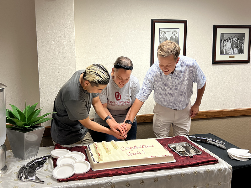 Three students standing behind a large cake, cutting the cake