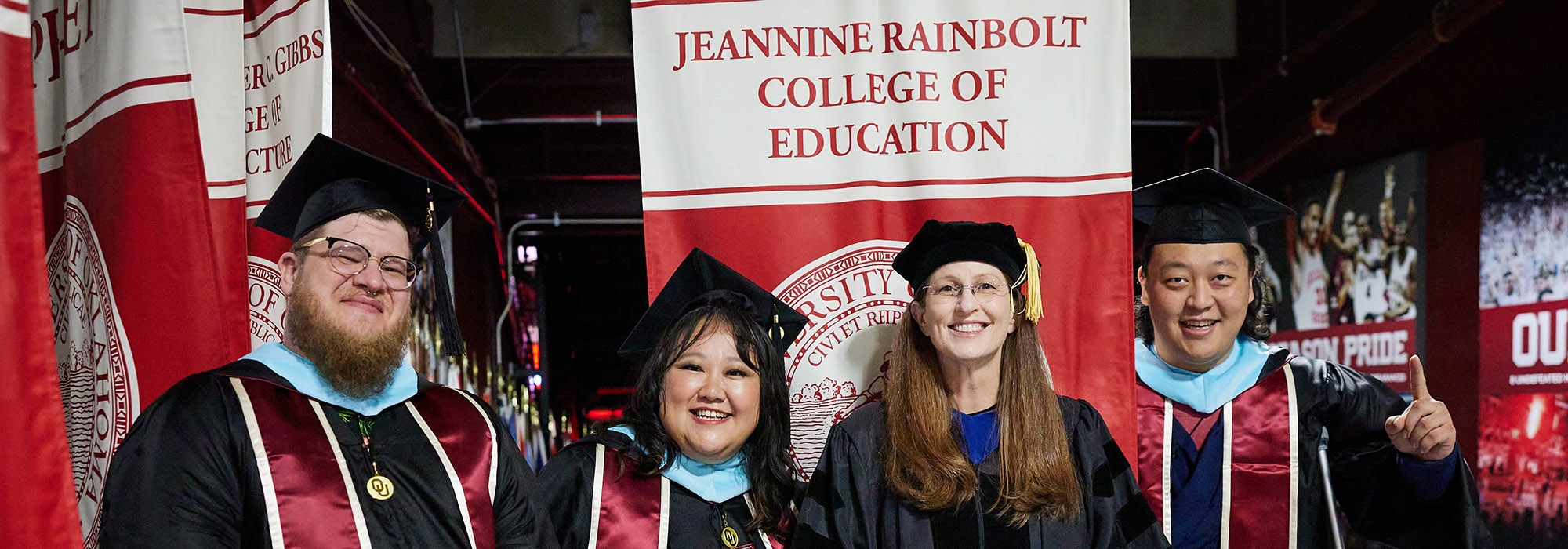 Students and faculty in graduation regalia standing in front of a Jeannine Rainbolt College of Education banner.