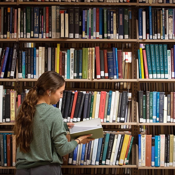 Student reading a book in front of a library's shelves.