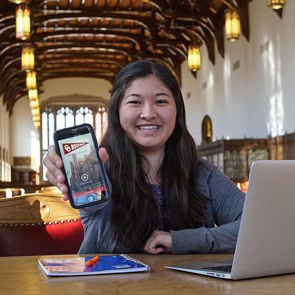 A student sitting in the Great Reading Room, holding up a phone.