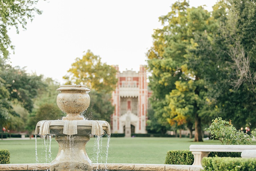 A water fountain encircled by white geraniums adorns the south oval of the University of Oklahoma campus in Norman, OK. A fine example of Cherokee Gothic architecture, the Bizzell Library, is featured in the background.
