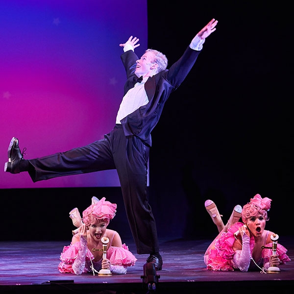 Two student performers in costume, talking on phones, with a student in a tuxedo dancing in the foreground.