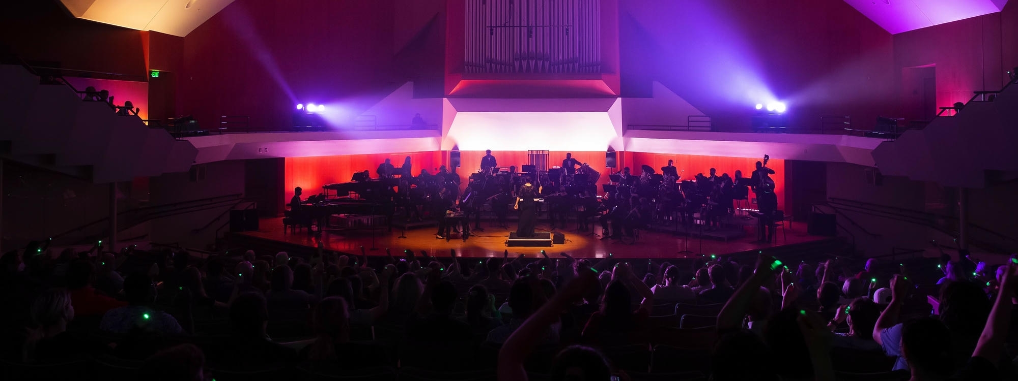 The OU Wind Symphony performs in Sharp Hall with lighting effects and a packed crowd. Some crowd members hold green lights, a part of the performance.