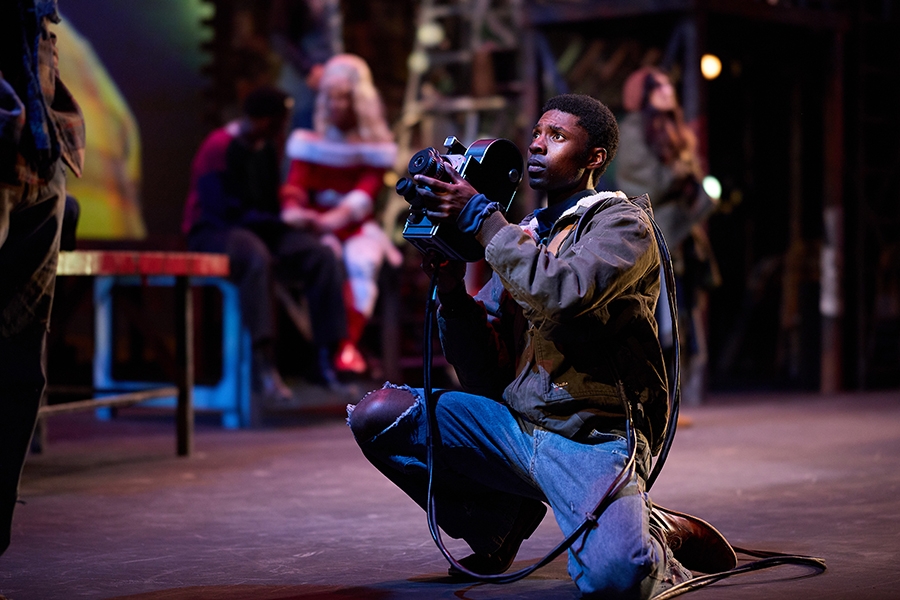 An actor in a production of RENT, kneeling down while holding a camera.