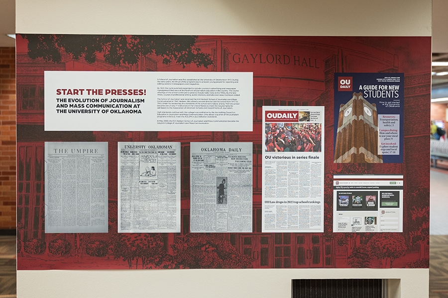 A large antique printing press, once kept in storage, now displayed as the centerpiece of the Gaylord College exhibit at OU’s Bizzell Library, surrounded by historical photographs and typewriters.