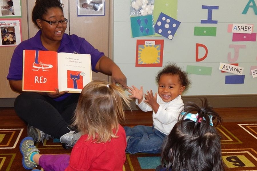 Picture of school teacher reading a book to two young children on the floor in classroom.
