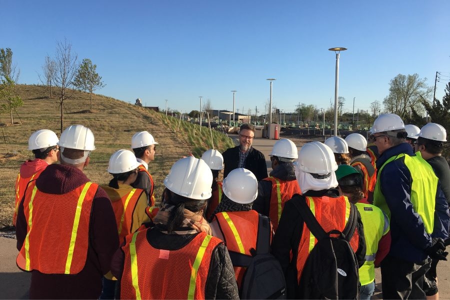 Picture of students in hardhats and vests looking at Gavin McMillan speak to them at O K C Scissortail Park.