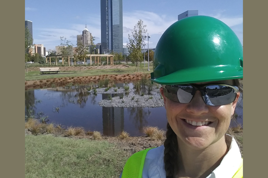 Selfie of alumna Kimberly White in a hard hat in front of distant skyscrapers of OKC